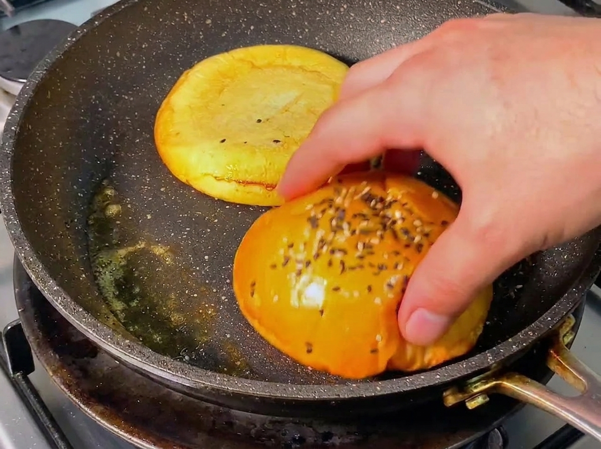Una mano che gira la metà di un panino da burger dorata in una padella ben calda con burro fuso.
