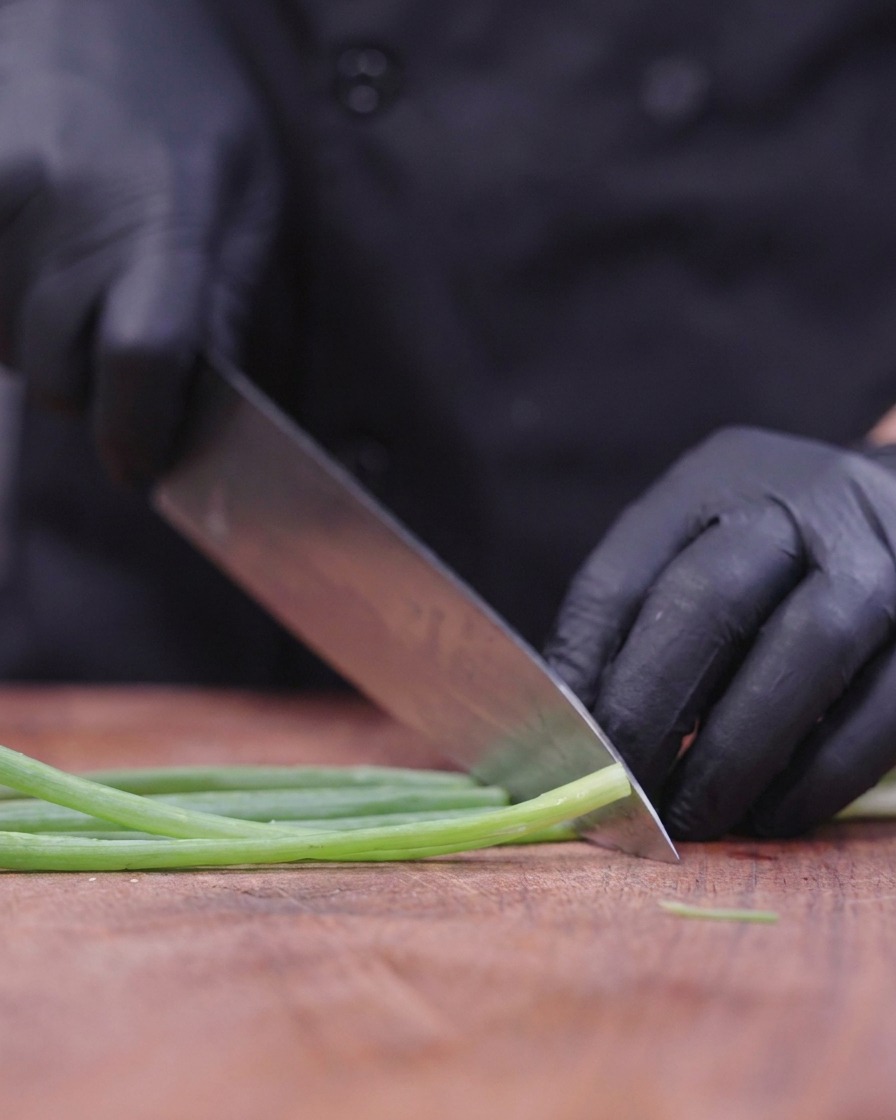 Uno chef con guanti neri usa un grande coltello per affettare finemente i cipollotti su un tagliere di legno.