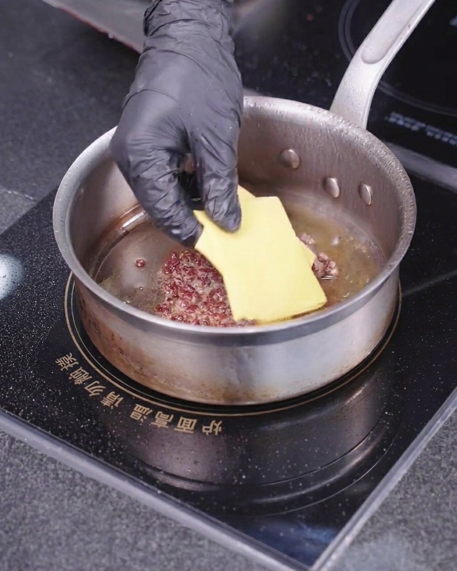 Una mano con guanto posiziona una fetta quadrata di formaggio giallo sopra un tortino di carne in cottura dentro una padella calda.