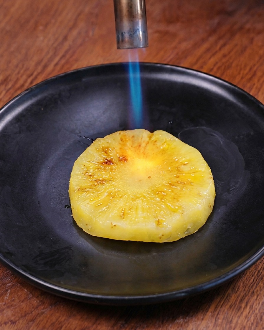 La fiamma blu di un cannello da cucina che dora la superficie di una fetta di ananas fresco su un piatto scuro.