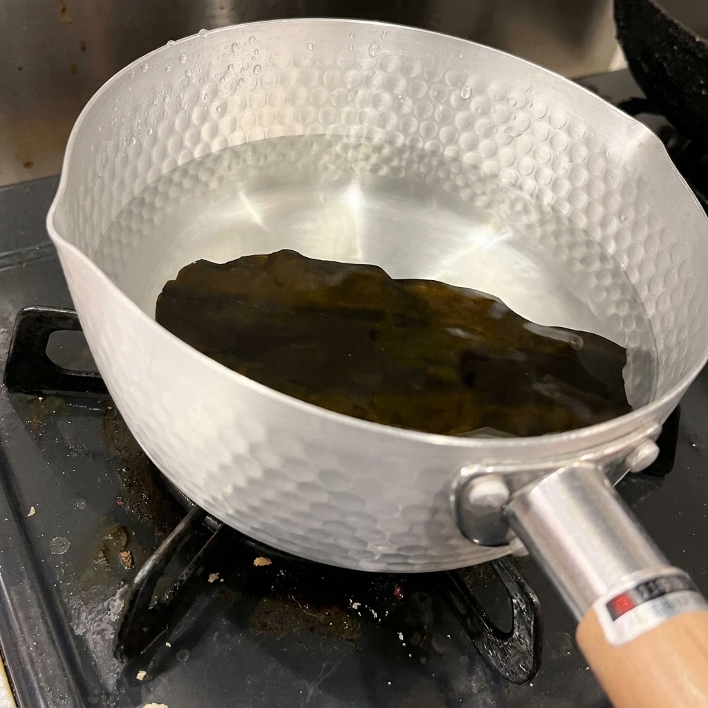 Un pezzo di alga kombu scura ed essiccata sul fondo di una pentola in alluminio battuto piena di acqua fredda e limpida.