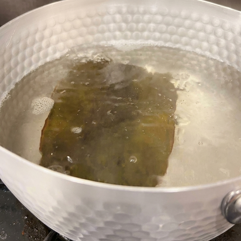 Lacqua freme leggermente attorno a un pezzo di alga kombu reidratata allinterno di una pentola argentata sul fuoco.