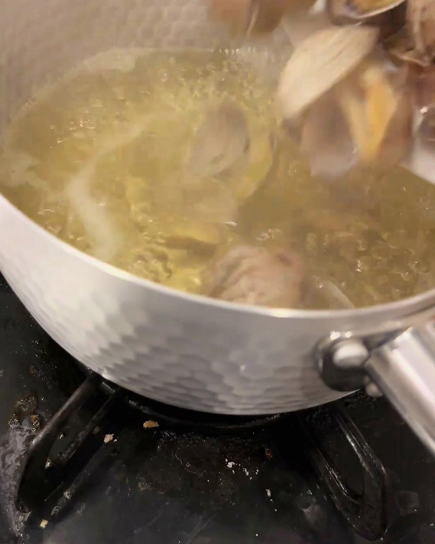 Vongole fresche tuffate in un dashi limpido a inizio ebollizione allinterno di una pentola di metallo su un fornello a gas.