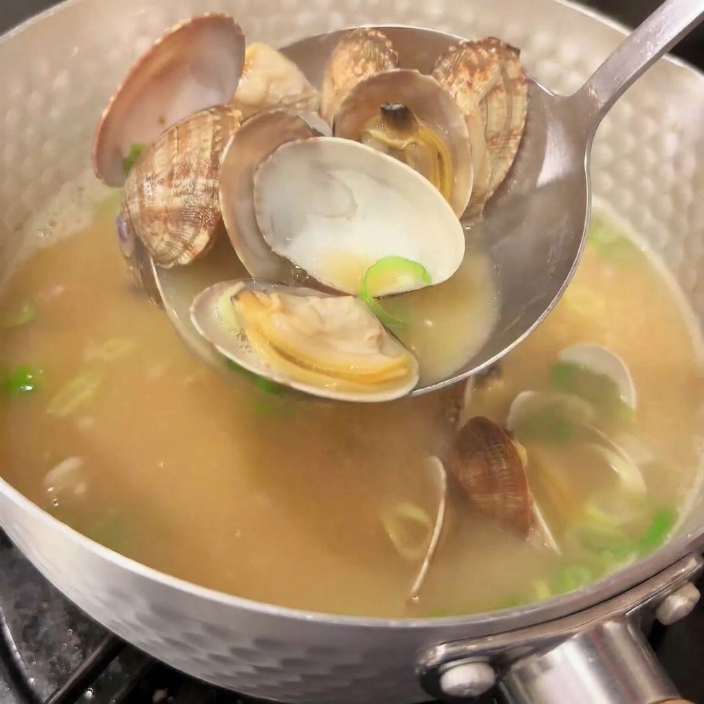Un mestolo di metallo raccoglie le vongole aperte da una pentola di zuppa di miso giapponese fumante, con una pioggia di cipollotto fresco tritato nel brodo.
