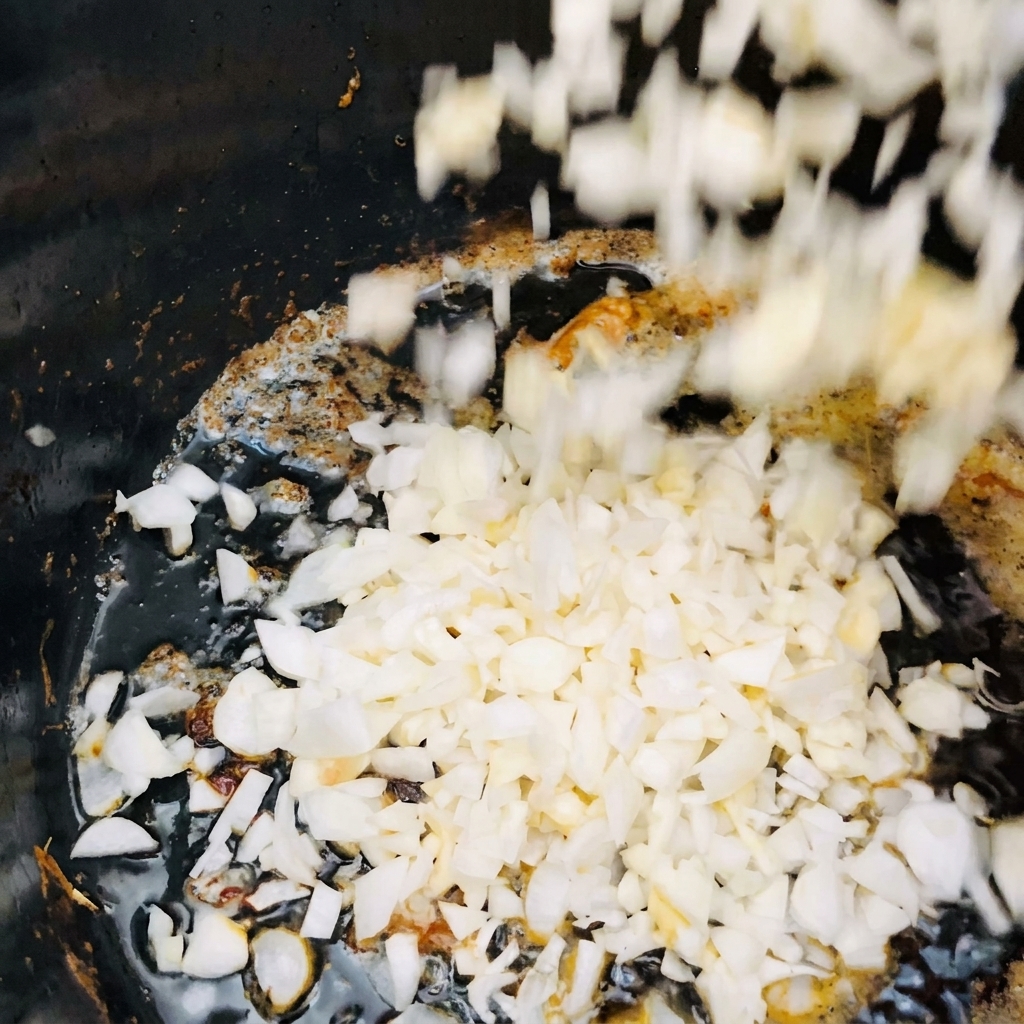 Cipolle bianche a dadini e aglio tritato che cadono in una pentola scura con olio caldo e brillante.
