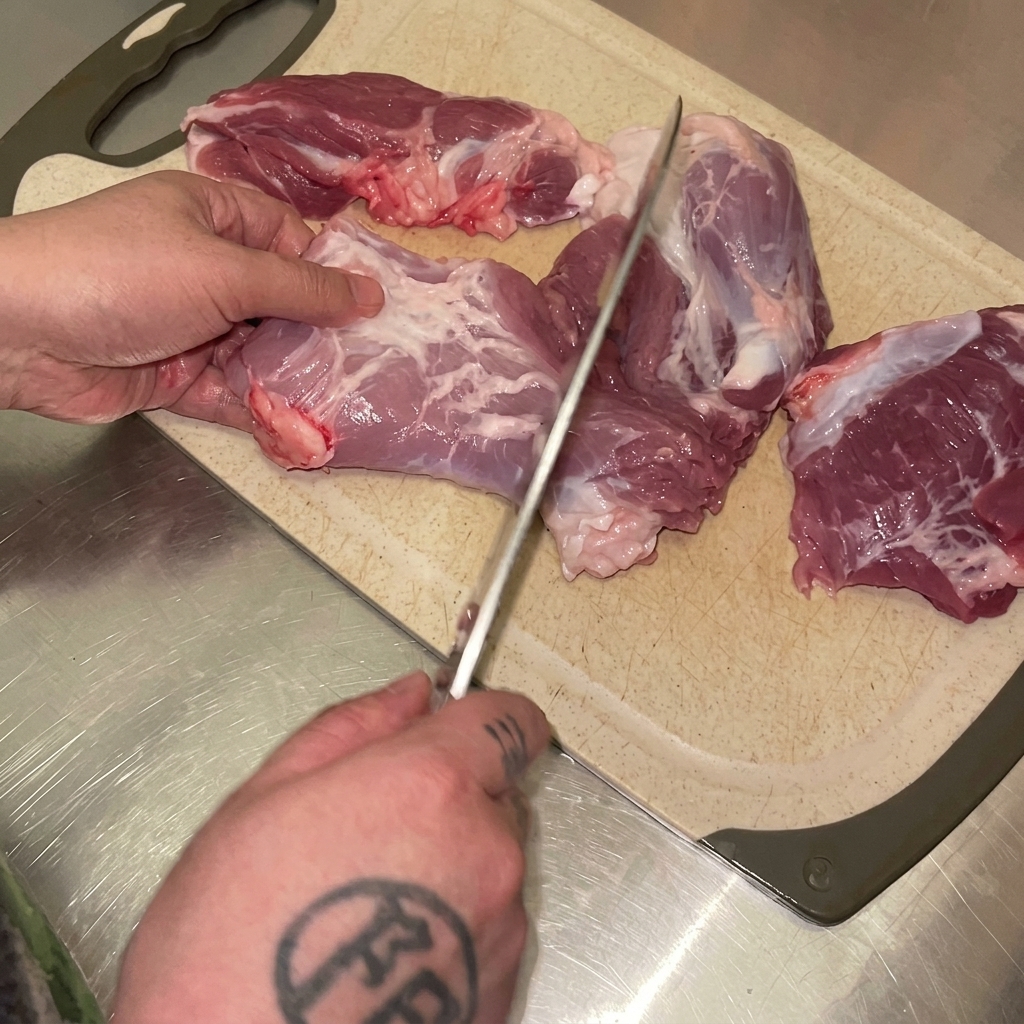 Mani che usano un coltello per tagliare la coscia di maiale cruda a cubetti su un tagliere.