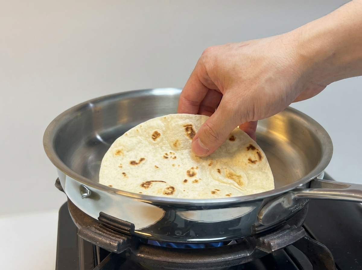 Una mano che posiziona una tortilla di farina soffice e spessa su una padella in acciaio inossidabile riscaldata.
