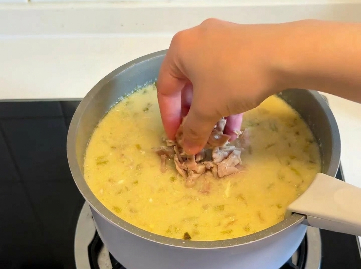 Una mano che aggiunge pezzi di pollo cotto e sfilacciato in una pentola di zuppa cremosa al limone.