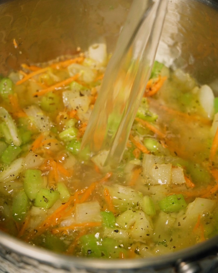 Brodo di pollo versato in una pentola contenente verdure saltate ed erbe aromatiche.