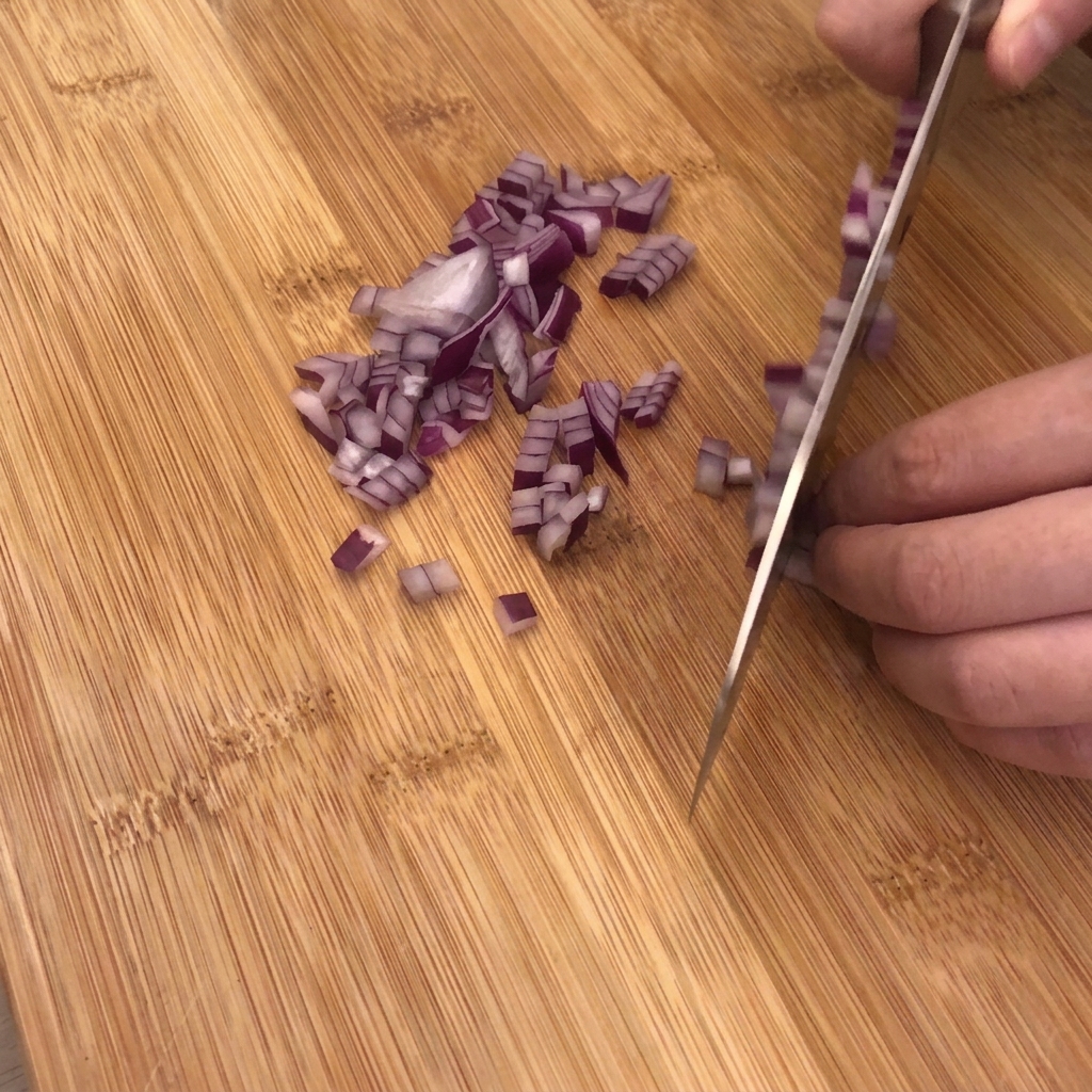 Un coltello da cucina affilato trita una cipolla rossa su un tagliere in legno con aglio e pezzi di pancetta nelle vicinanze.
