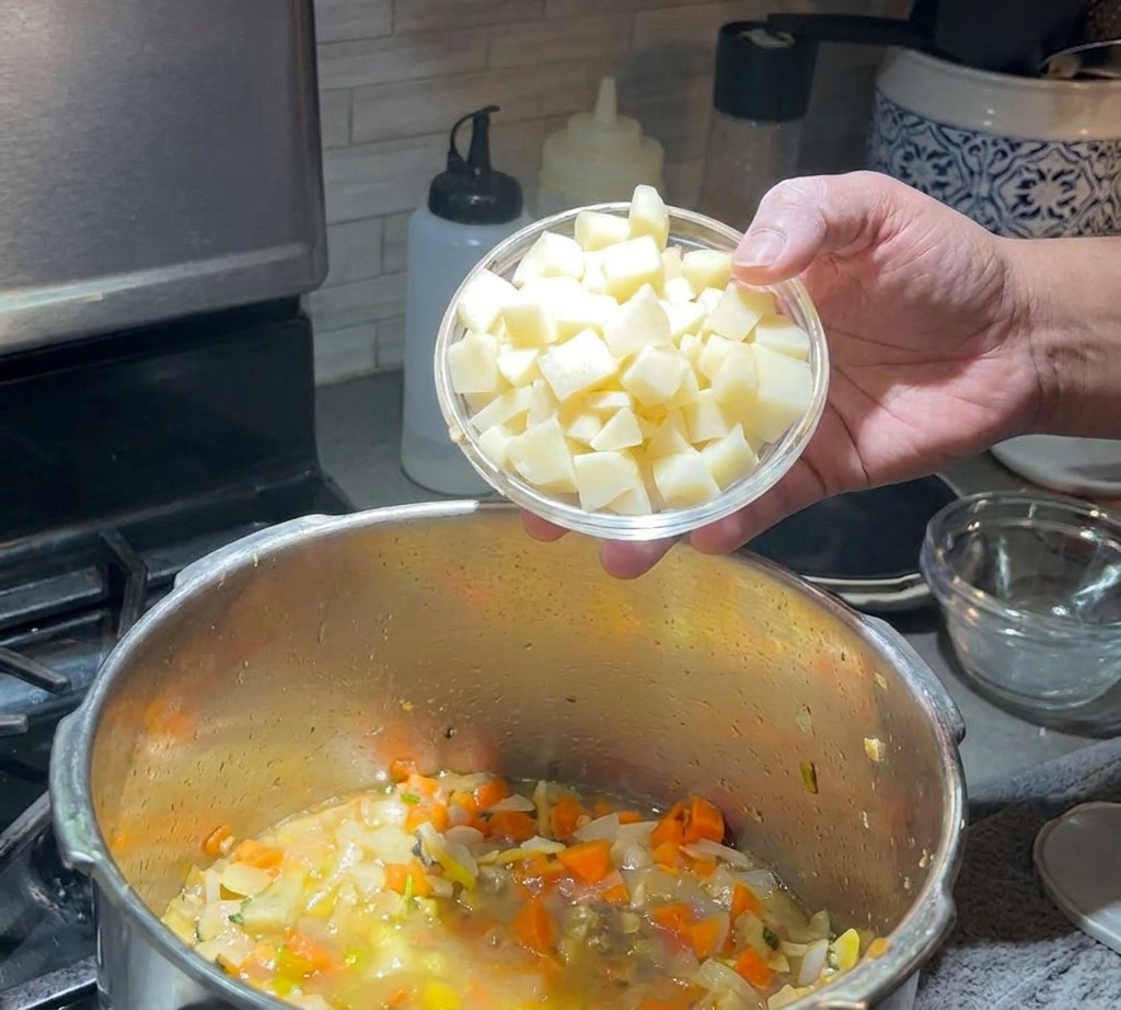 Una mano che versa una piccola ciotola di patate a cubetti in una pentola di zuppa in ebollizione con carote e cipolle.