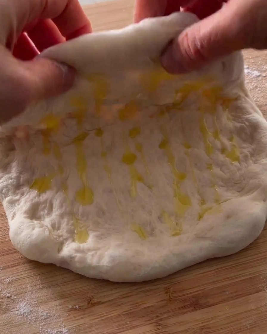 Mani che piegano a metà un pezzo di impasto della pizza oliato su una superficie di legno.