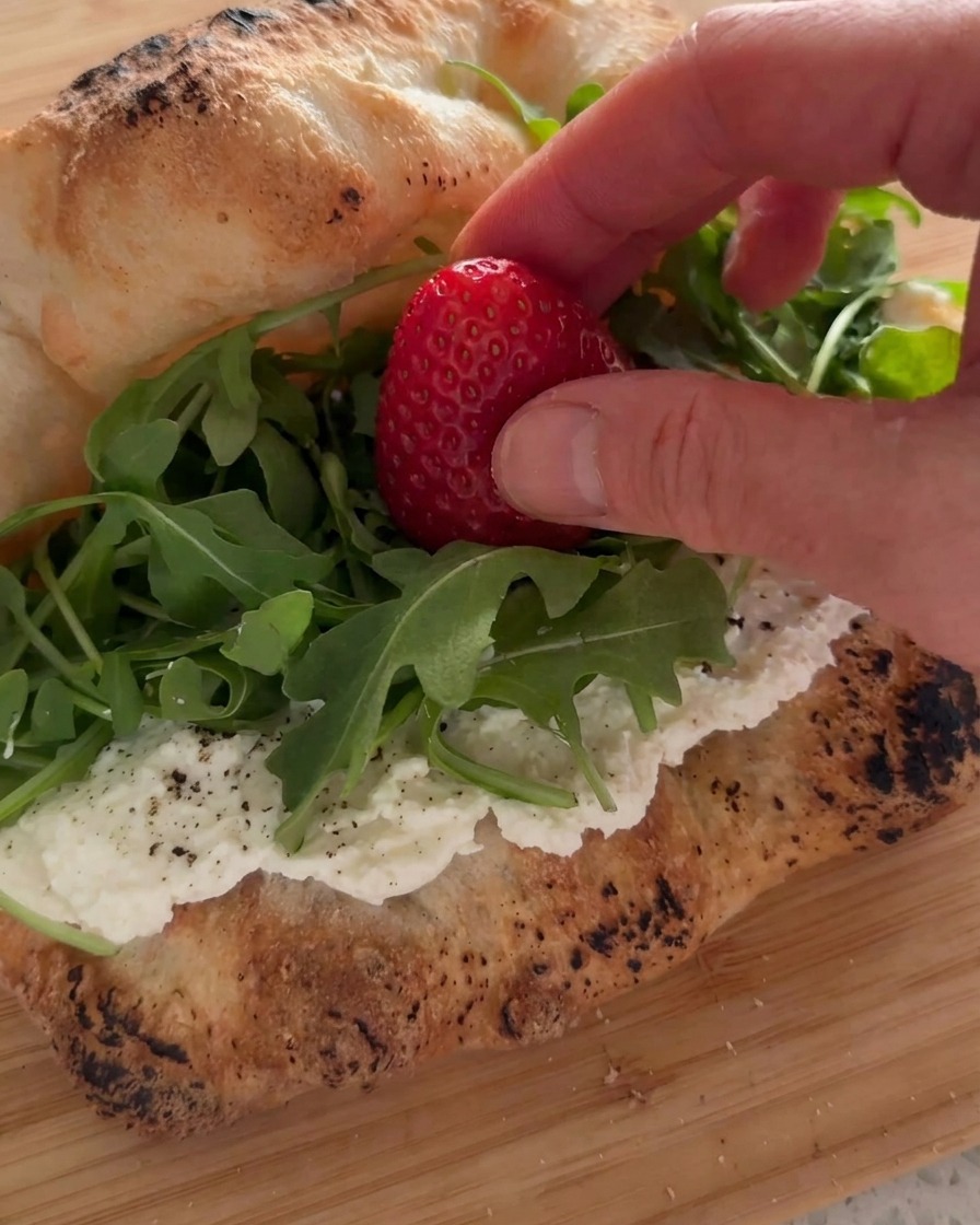 Una mano posiziona una fragola fresca su un letto di rucola allinterno di un panino con uno strato di ricotta.
