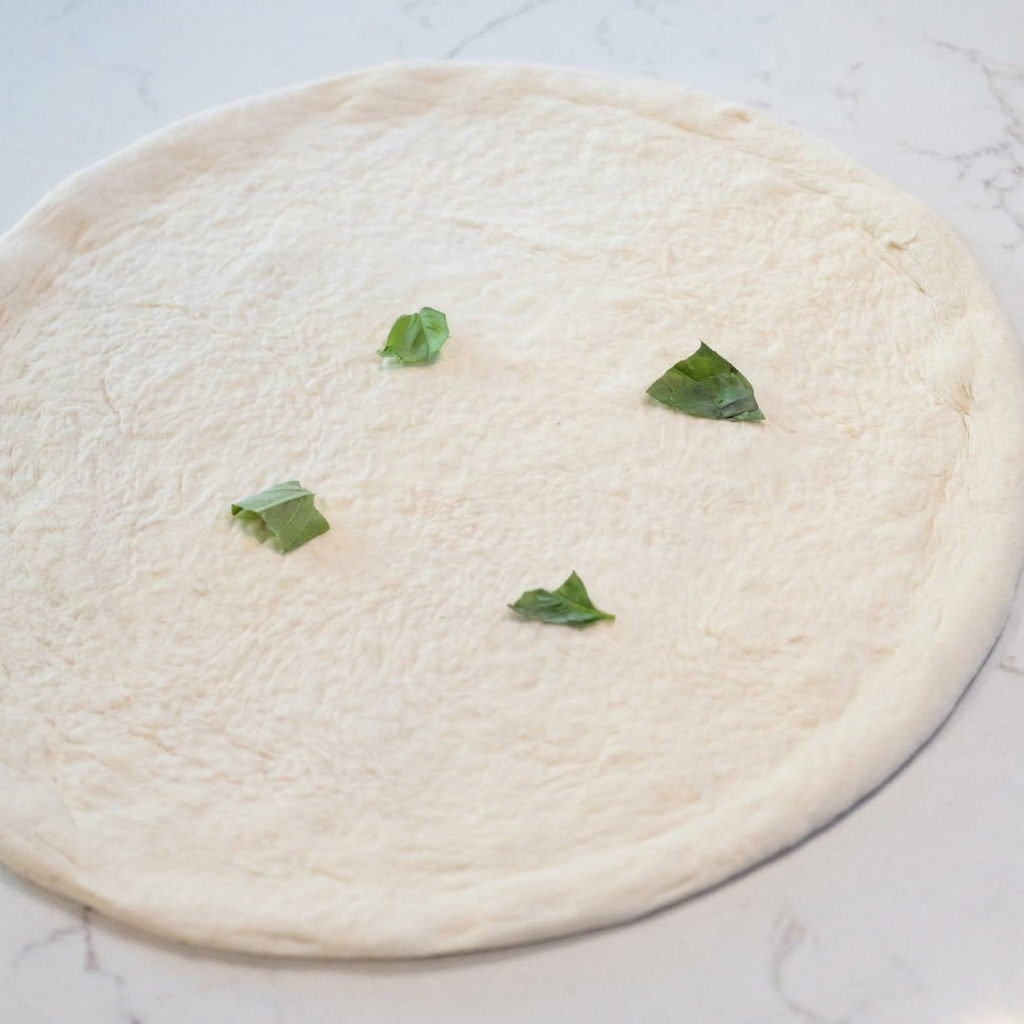 Un disco di impasto crudo per pizza guarnito con foglie verdi e fresche di basilico sparse sulla superficie.