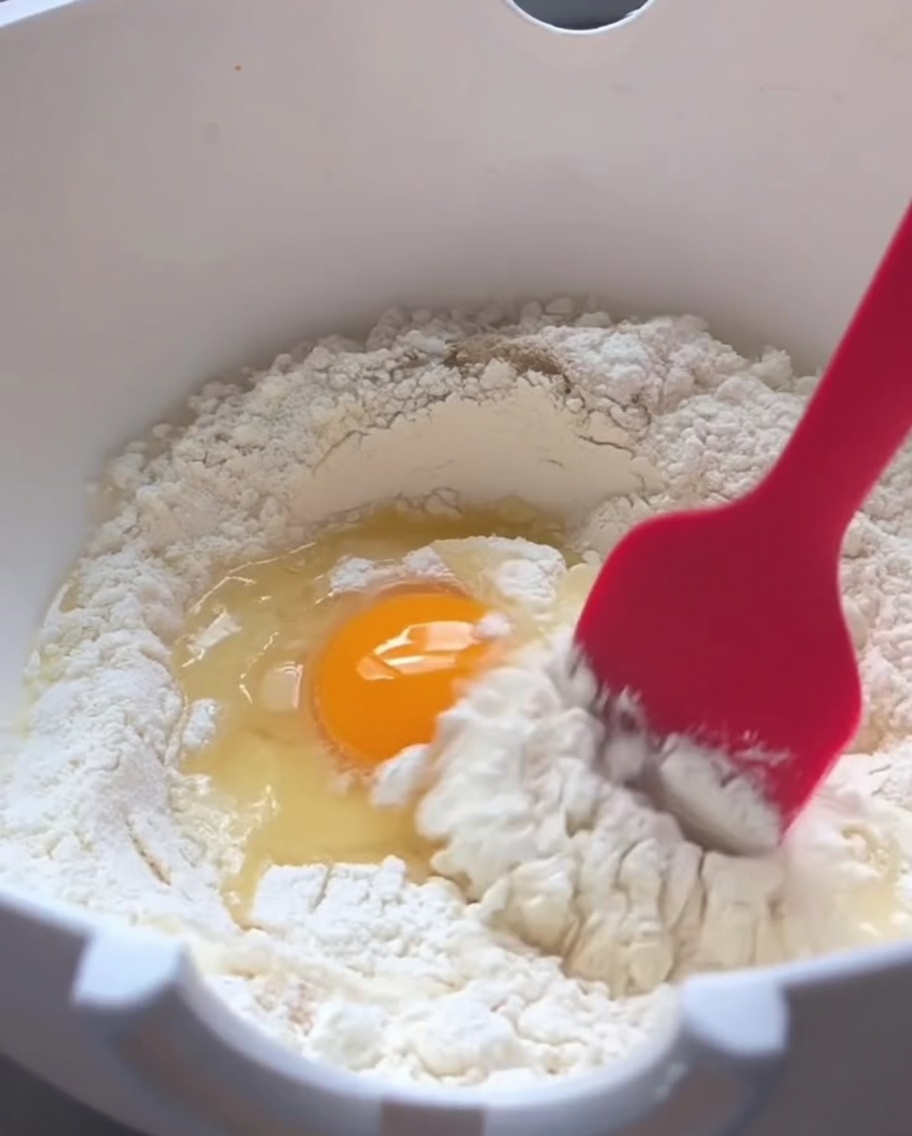 Una spatola in silicone rossa mescola farina, un uovo crudo e acqua tiepida in una ciotola bianca.