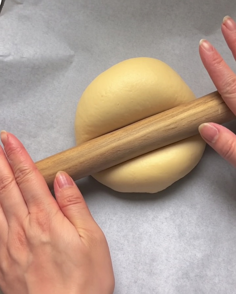 Mani che usano un mattarello di legno per appiattire la pasta su carta forno.