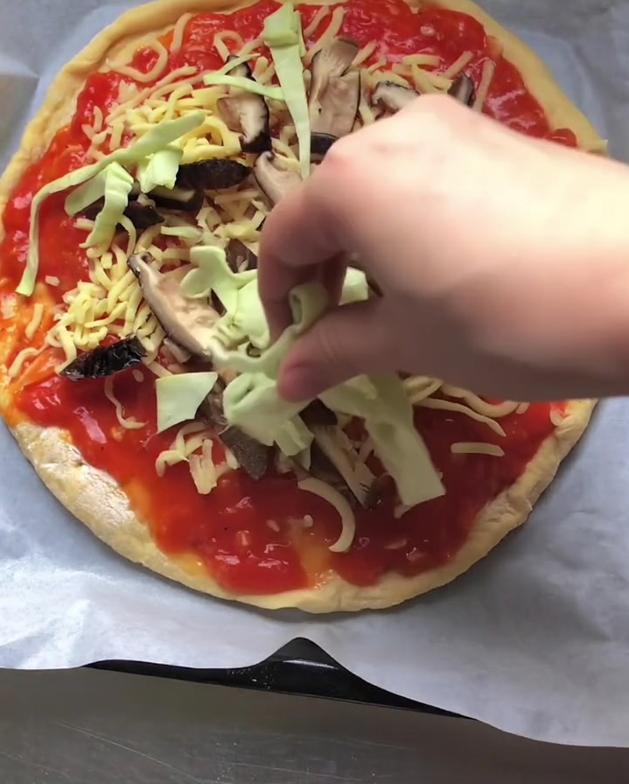 Mano che distribuisce cavolo verza tritato sopra una pizza con sugo, formaggio e funghi.