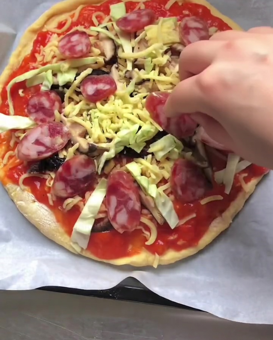 Mano che posiziona fettine di salsiccia su una pizza cruda con verdure e formaggio.