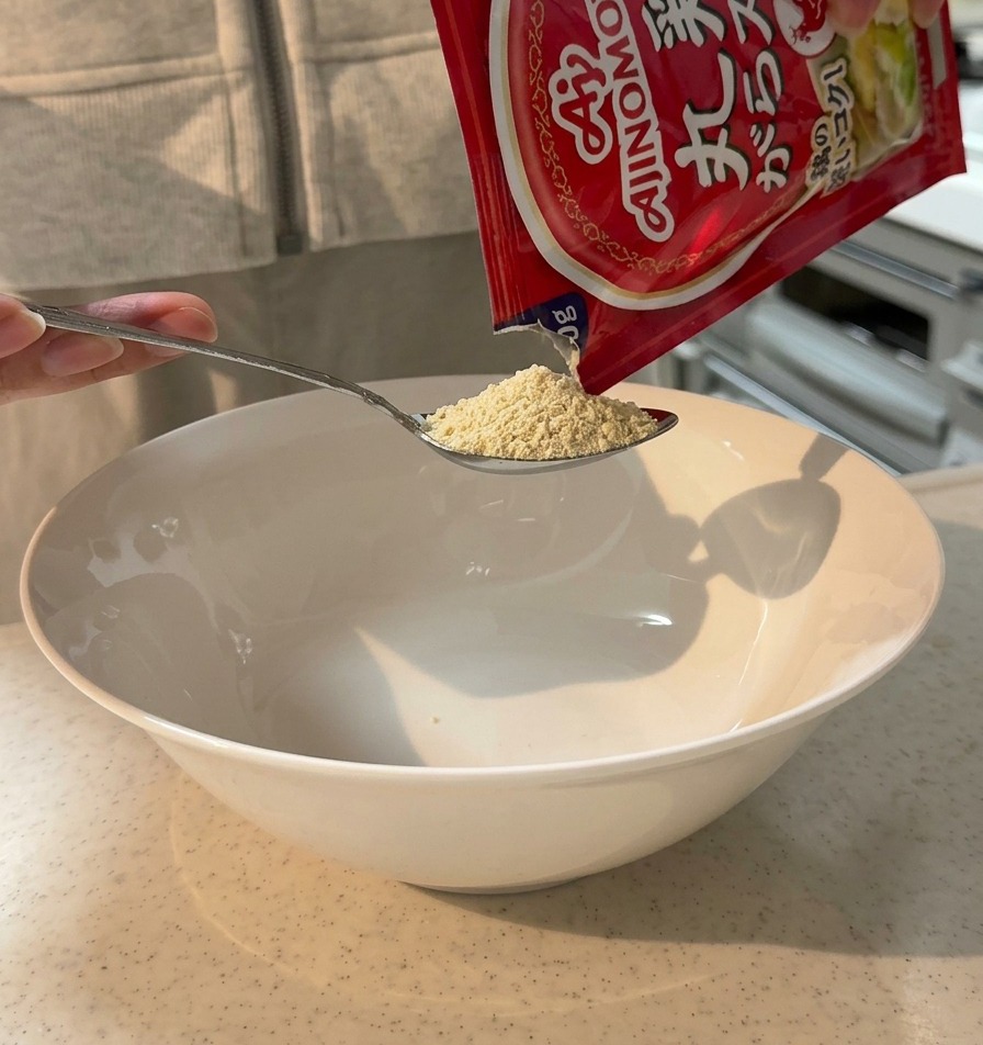 Preparato per brodo di pollo in polvere versato da una bustina su un cucchiaio sopra una grande ciotola bianca.