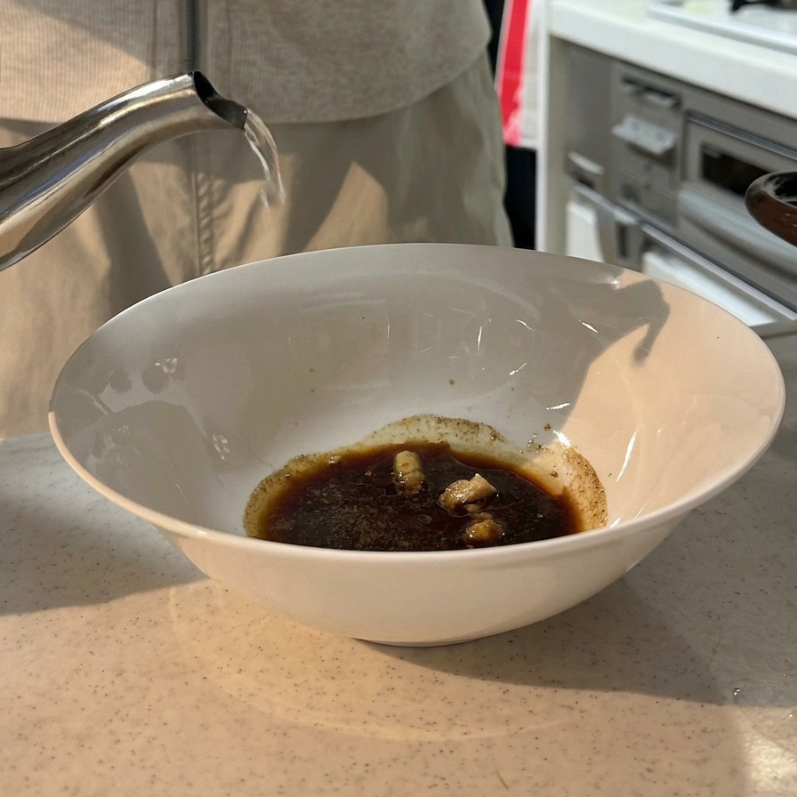Acqua bollente versata da un bollitore in acciaio inossidabile in una ciotola bianca contenente un mix di salsa di soia scura e spezie.