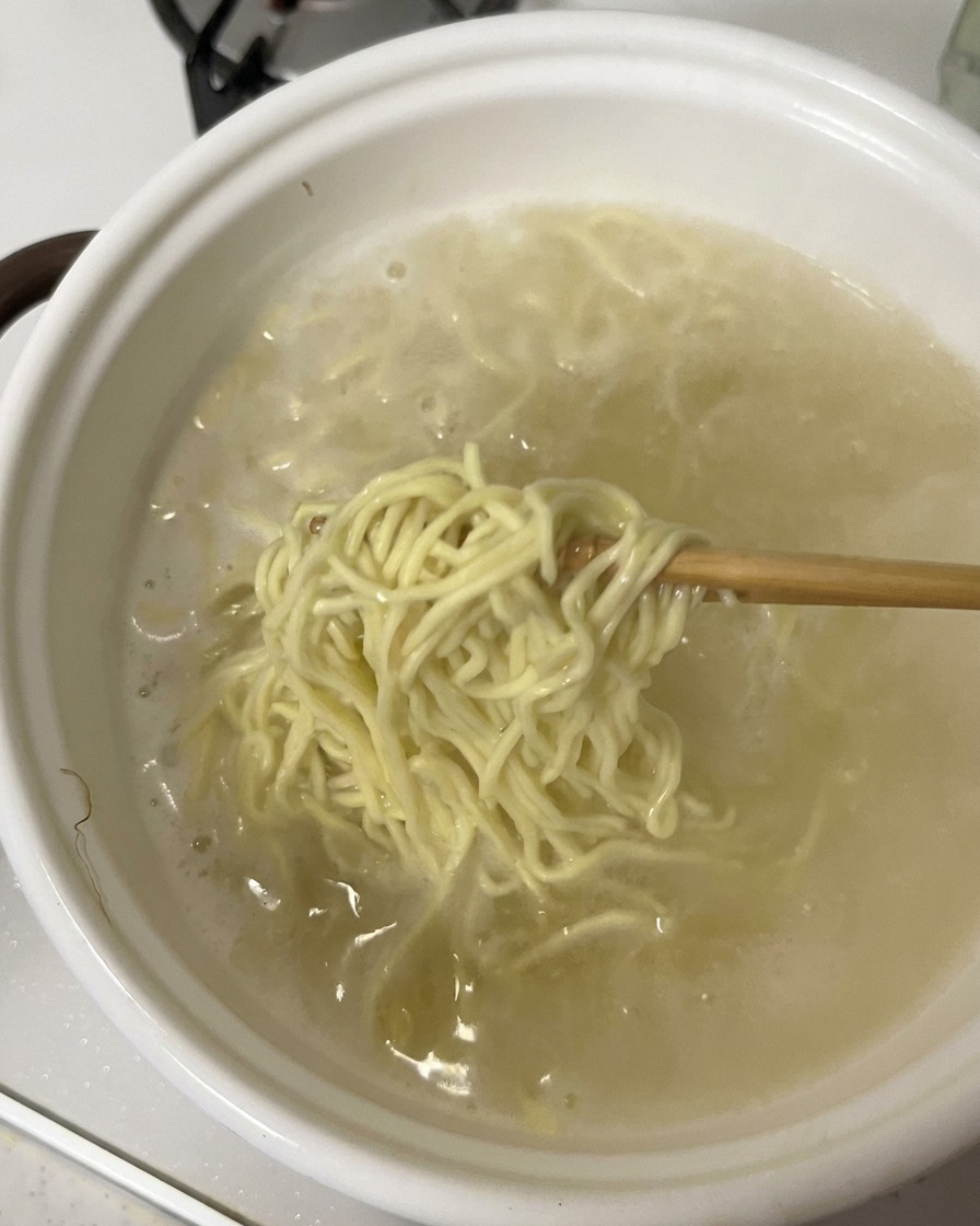 Bacchette che sollevano una porzione di noodles ramen cotti da una pentola bianca piena di acqua bollente.