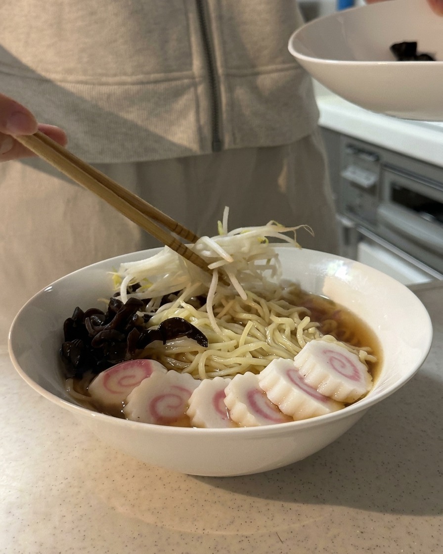 Bacchette che posizionano un mucchietto di germogli di soia sbollentati su una ciotola di ramen già guarnita con funghi e fette di surimi.