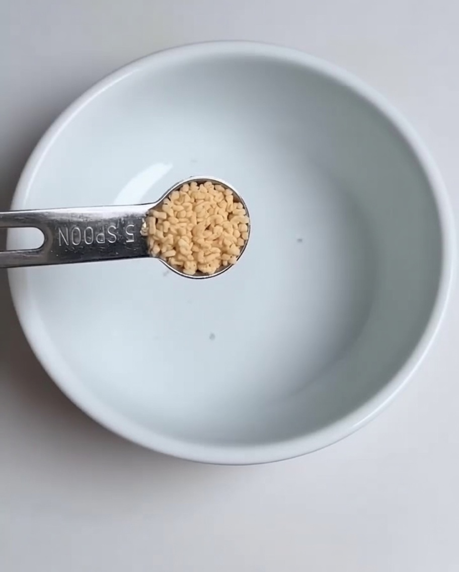 Un primo piano di un cucchiaino che aggiunge brodo di pollo in polvere in una ciotola bianca.