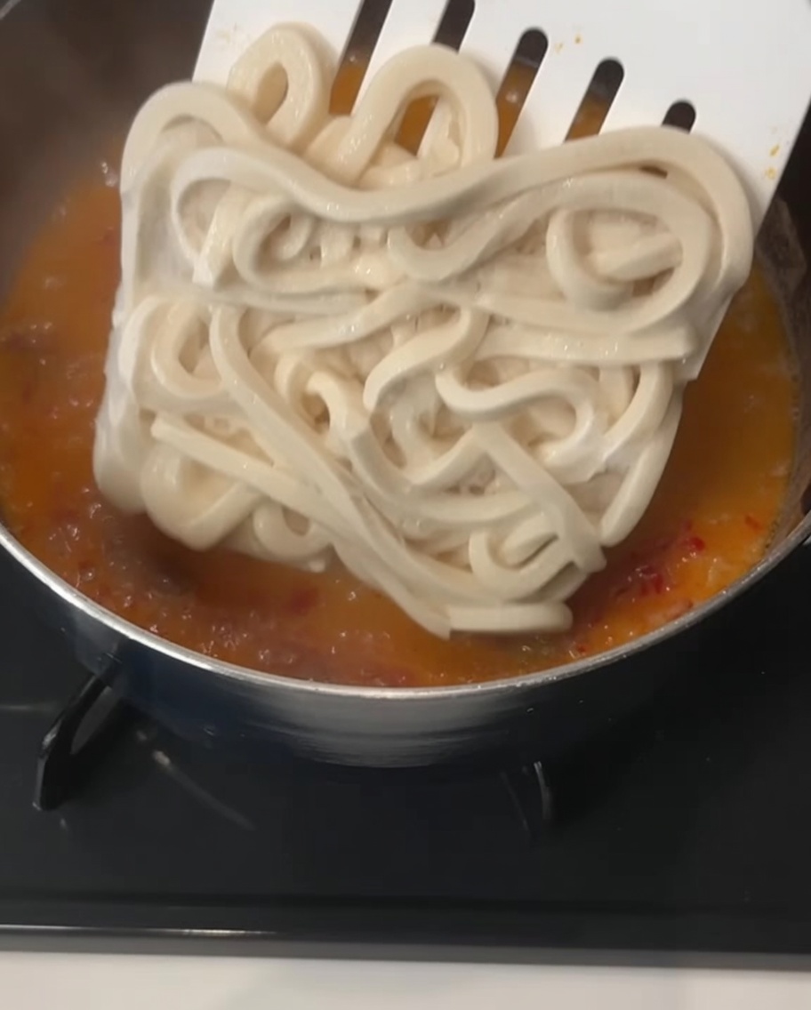 Un blocco quadrato di spessi noodle udon surgelati immerso con una spatola in un brodo arancione cremoso che sobbolle.