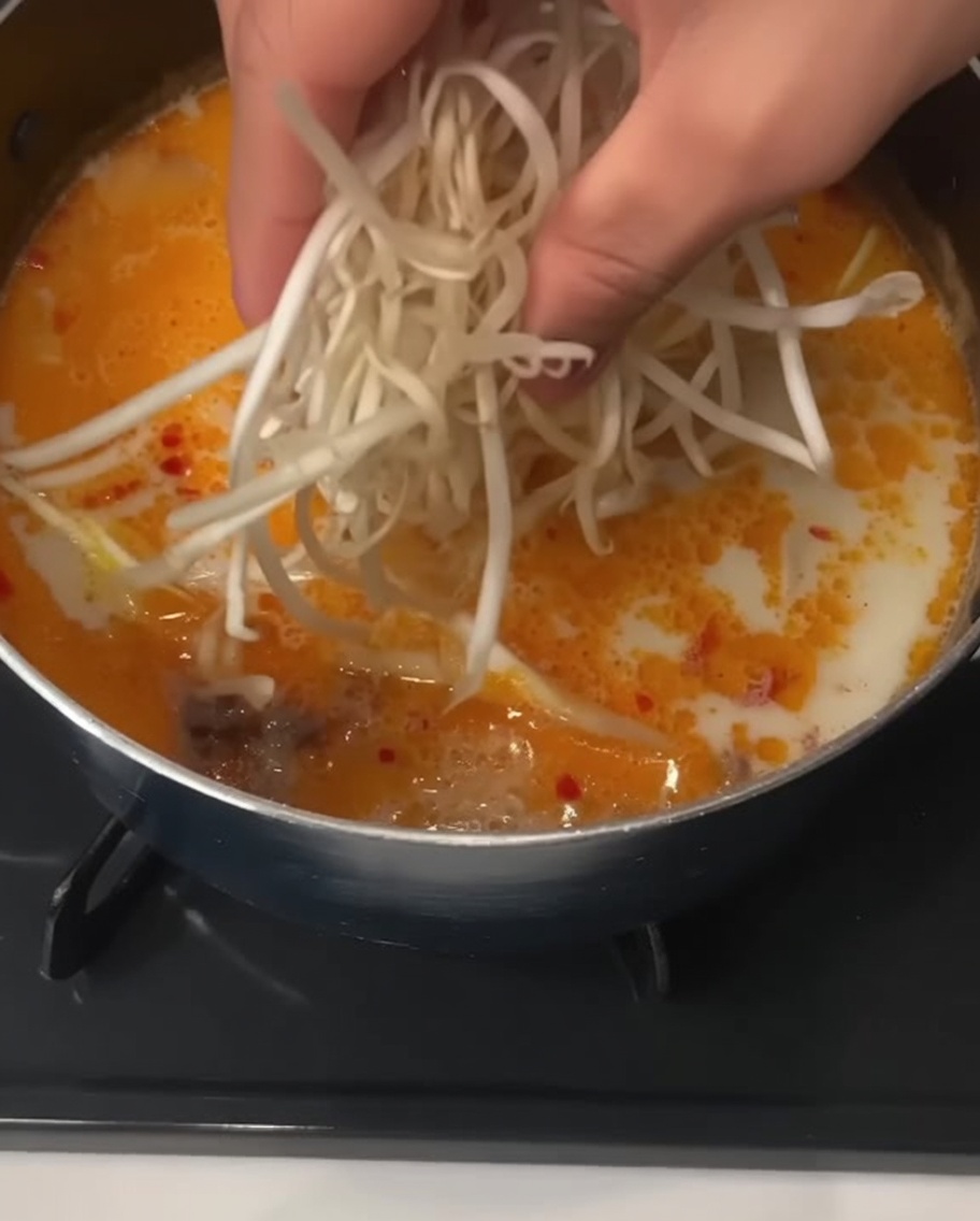 Una mano che aggiunge una manciata di germogli di soia freschi in una pentola di zuppa cremosa con noodle.