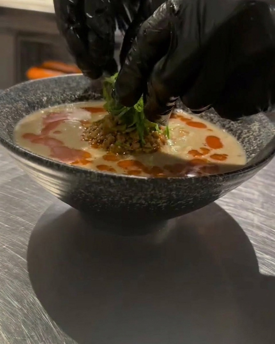 Mani guantate che posizionano con cura cipollotti affettati sopra una ciotola di Tan Tan Ramen caldo.