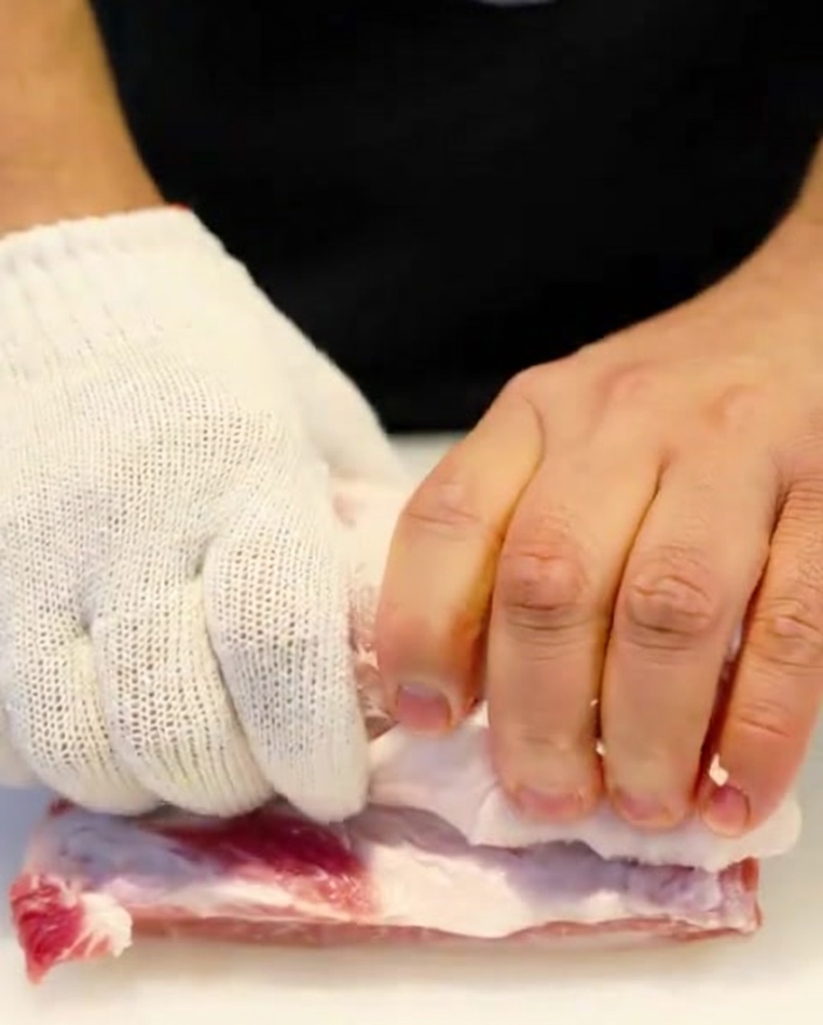 Un rotolo di pancetta di maiale cruda viene legato strettamente con dello spago da cucina da mani con guanti protettivi.