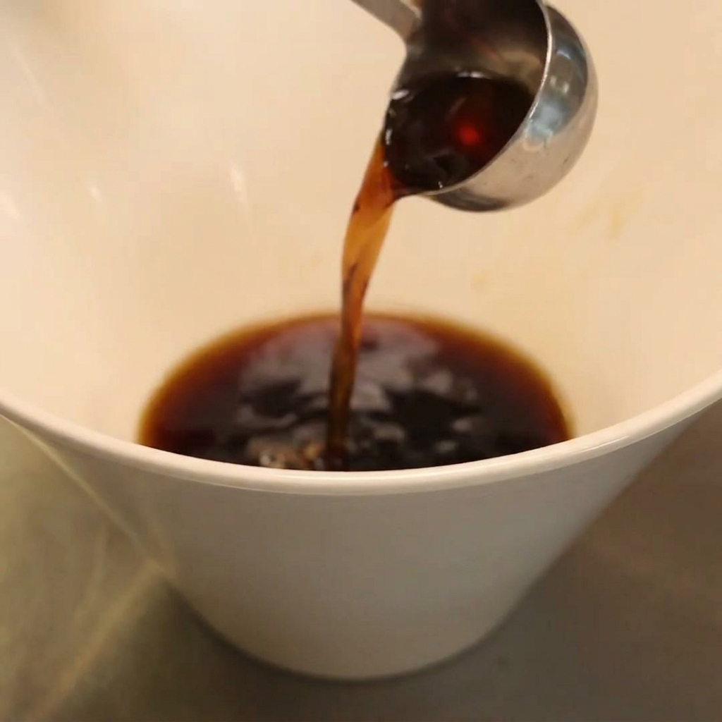 Condimento liquido scuro versato da un mestolo di metallo in una profonda ciotola bianca.