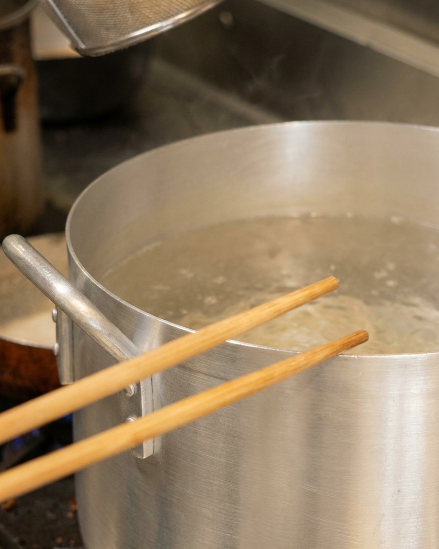 Bacchette di legno appoggiate su una grande pentola di alluminio con acqua bollente contenente un cestello per noodle.