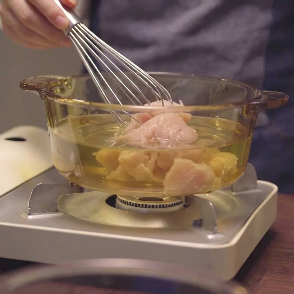 Una frusta che mescola petto di pollo macinato nel liquido allinterno di una pentola di vetro.