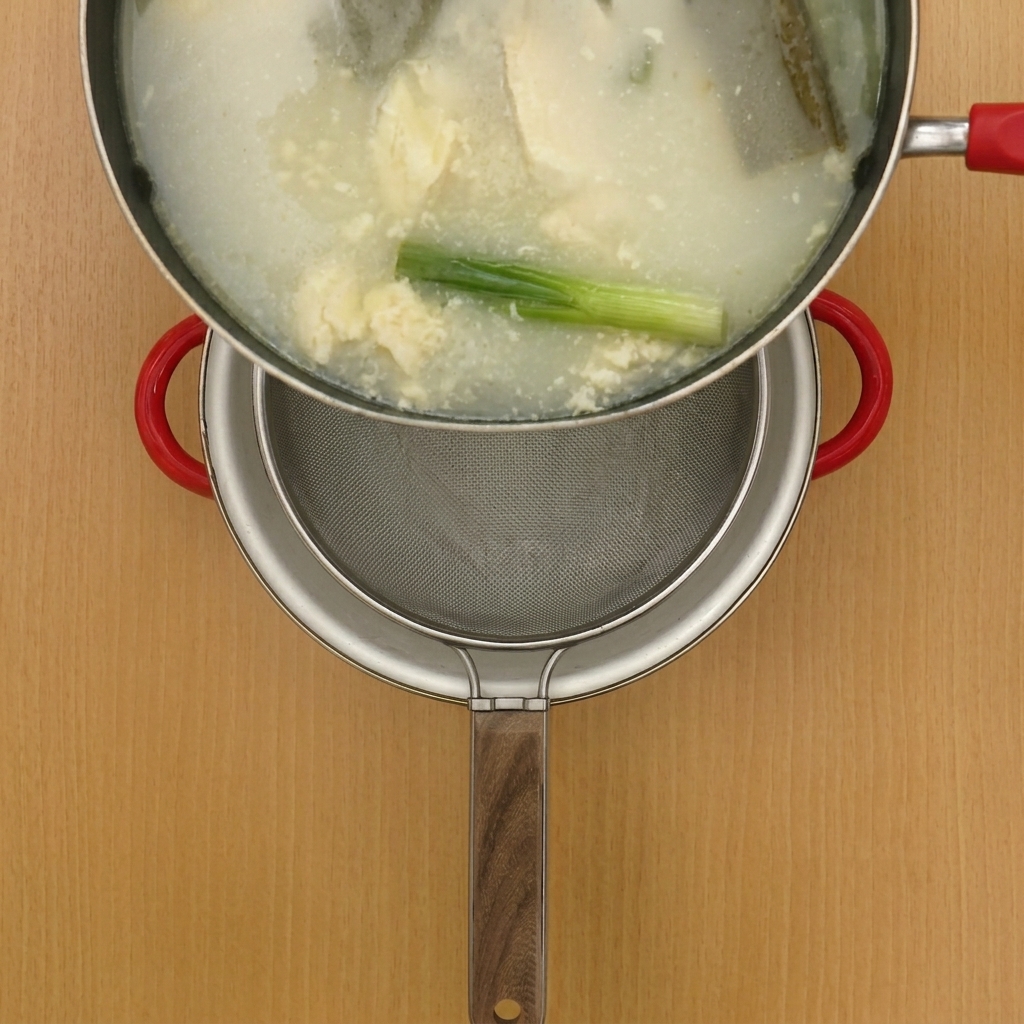 Brodo di pollo caldo versato attraverso un setaccio metallico in una pentola pulita.