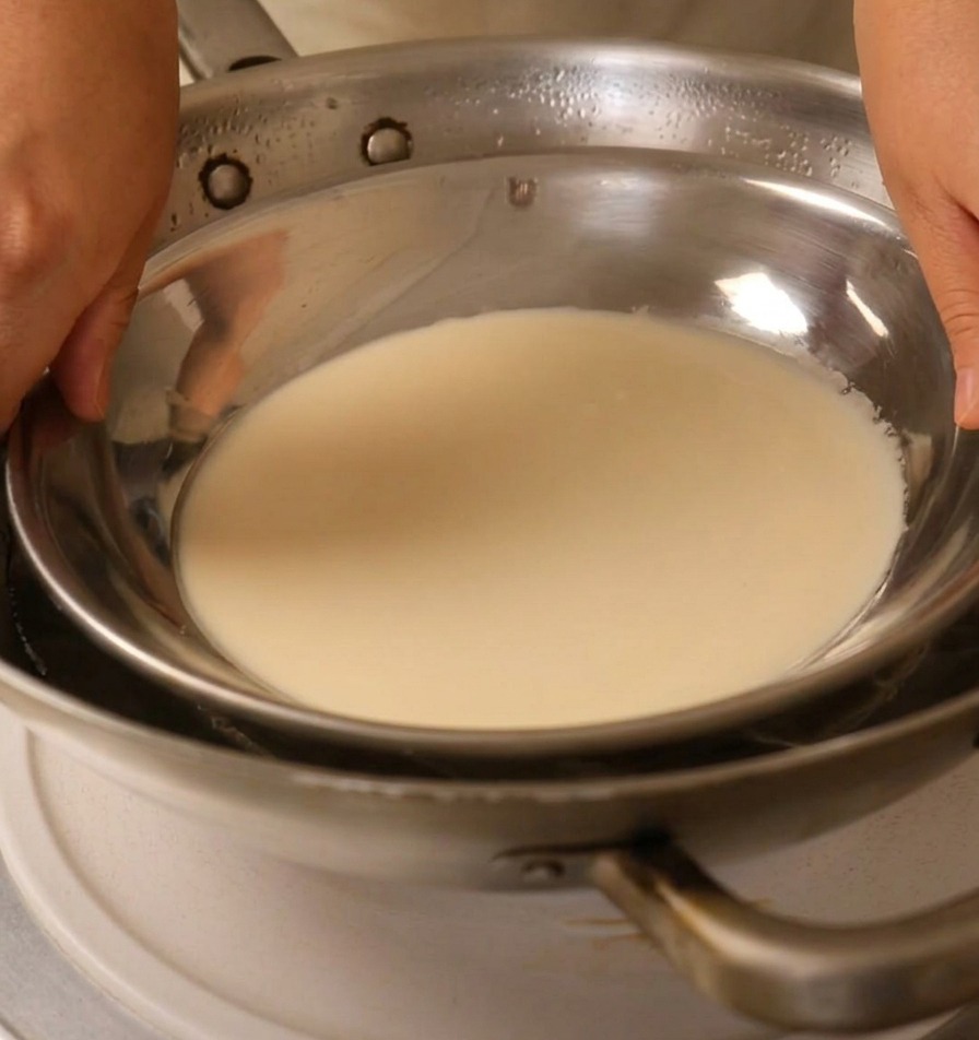 Mani che posizionano un piatto di metallo pieno di pastella bianca su un wok con acqua bollente.