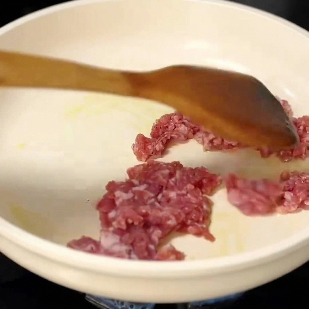 Maiale macinato crudo spezzettato e saltato in una padella bianca con una spatola di legno.