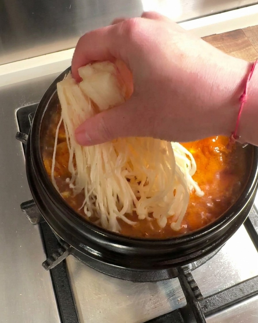 Funghi enoki freschi e fettine di manzo crudo inseriti nello stufato di kimchi in ebollizione.