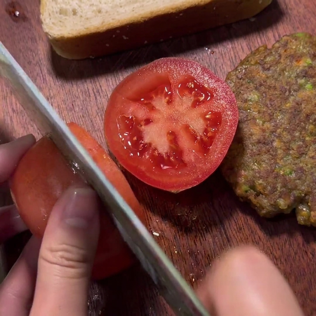 Una mano che affetta un pomodoro rosso fresco su un tagliere di legno accanto a un hamburger cotto.