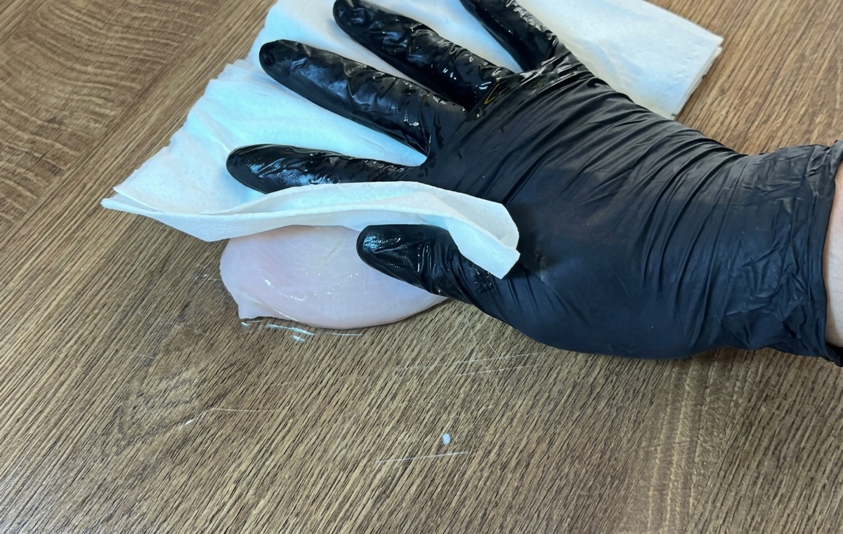 Una mano con guanto che usa un tovagliolo di carta per asciugare un petto di pollo crudo su una superficie di legno.