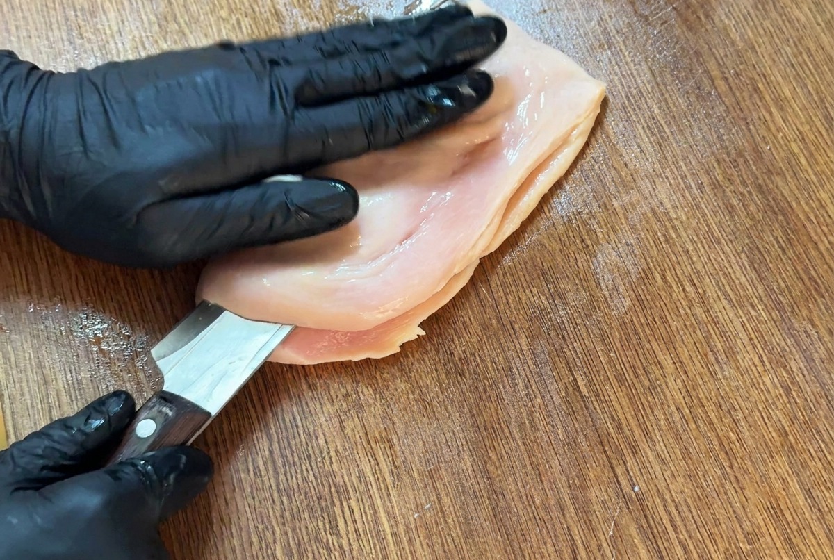 Un coltello affilato che taglia orizzontalmente un petto di pollo su un tagliere per aprirlo a libro.