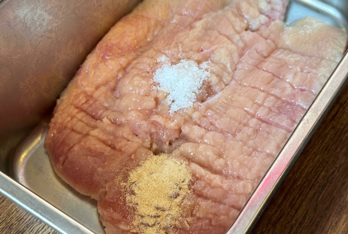 Un petto di pollo crudo in una teglia metallica ricoperto da varie spezie in polvere e salse.
