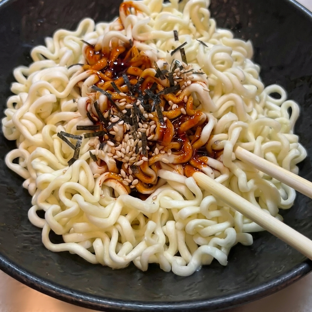 Noodles ramen ondulati in una ciotola nera strutturata mentre vengono mescolati con salsa piccante rossa, semi di sesamo e alga nori usando bacchette di legno.