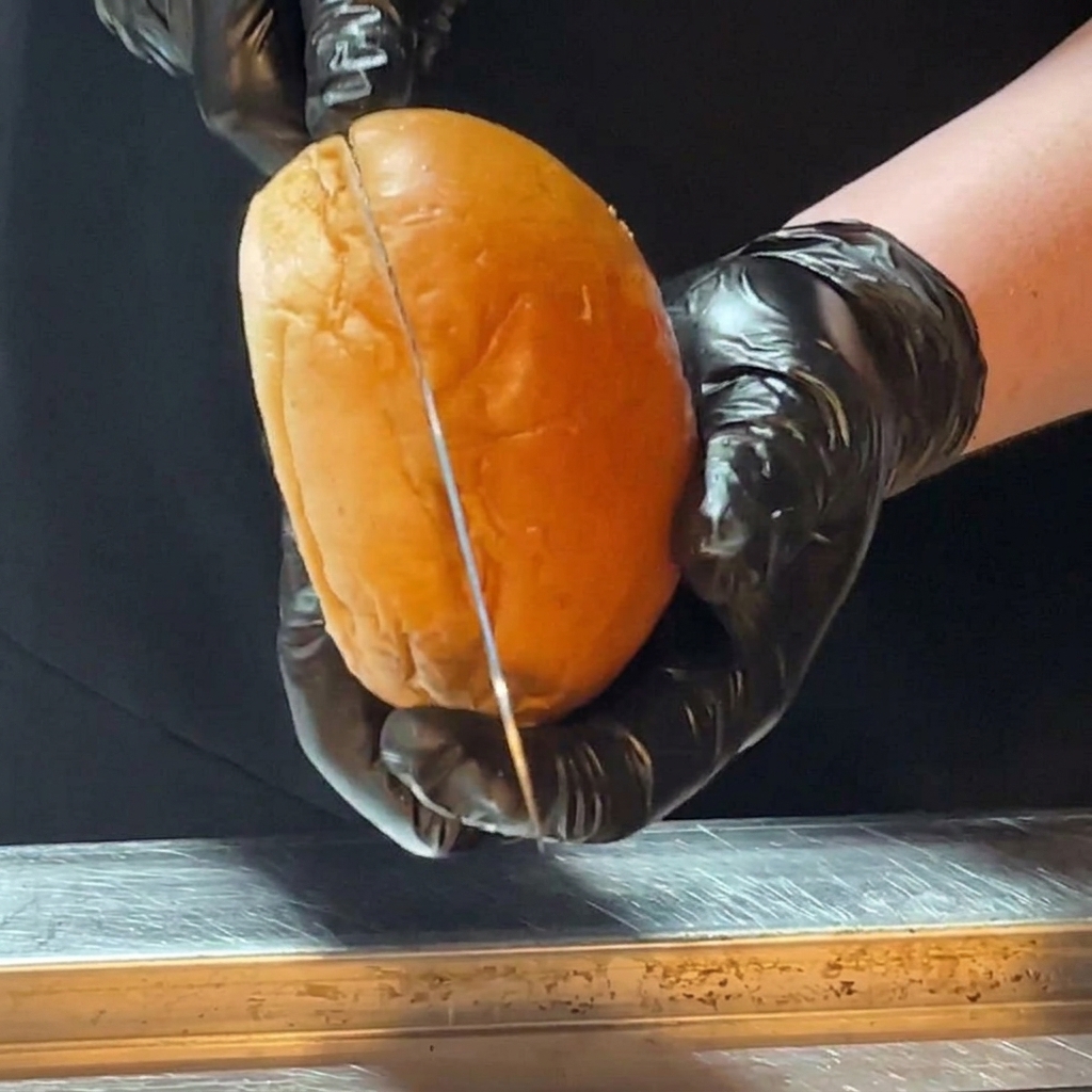 Mani con guanti neri da cucina usano un coltello lungo per tagliare a metà un lucido panino brioche.
