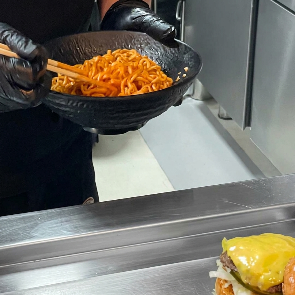Un burger al ramen piccante completato con un abbondante strato di noodles piccanti sopra il burger, pronto per essere chiuso con il panino superiore.
