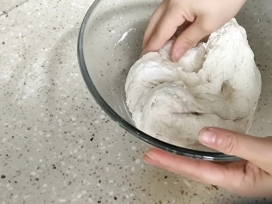Mani che premono e impastano una palla di pasta grezza allinterno di una ciotola di vetro.