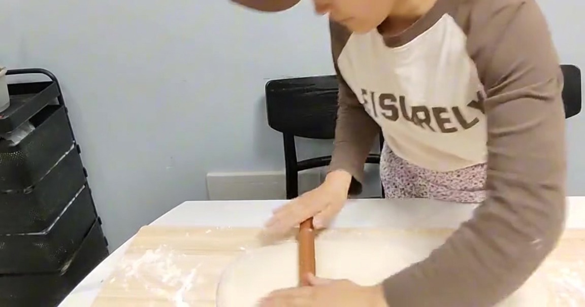Una persona usa un mattarello di legno per appiattire un blocco di pasta su una tavola di legno infarinata.