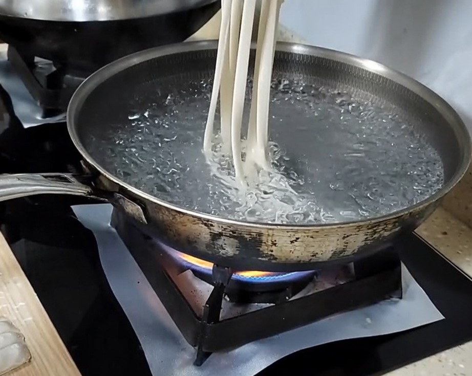 Mano che tira una striscia di pasta in un lungo noodle prima di lasciarlo cadere in una pentola di acqua bollente.