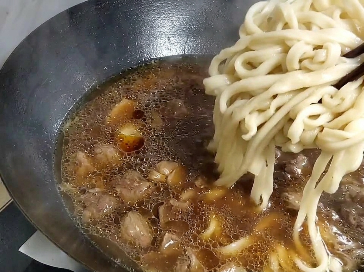 Noodles spessi sollevati con le bacchette da una pentola e messi in un wok con la base di zuppa.
