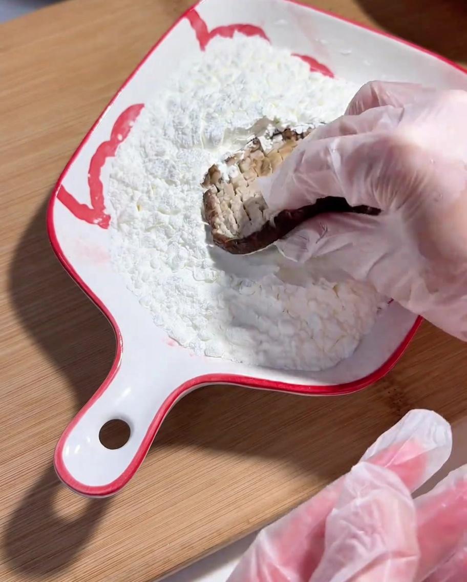 Una mano con guanto che intinge il lato inciso di un fungo shiitake nellamido di mais.
